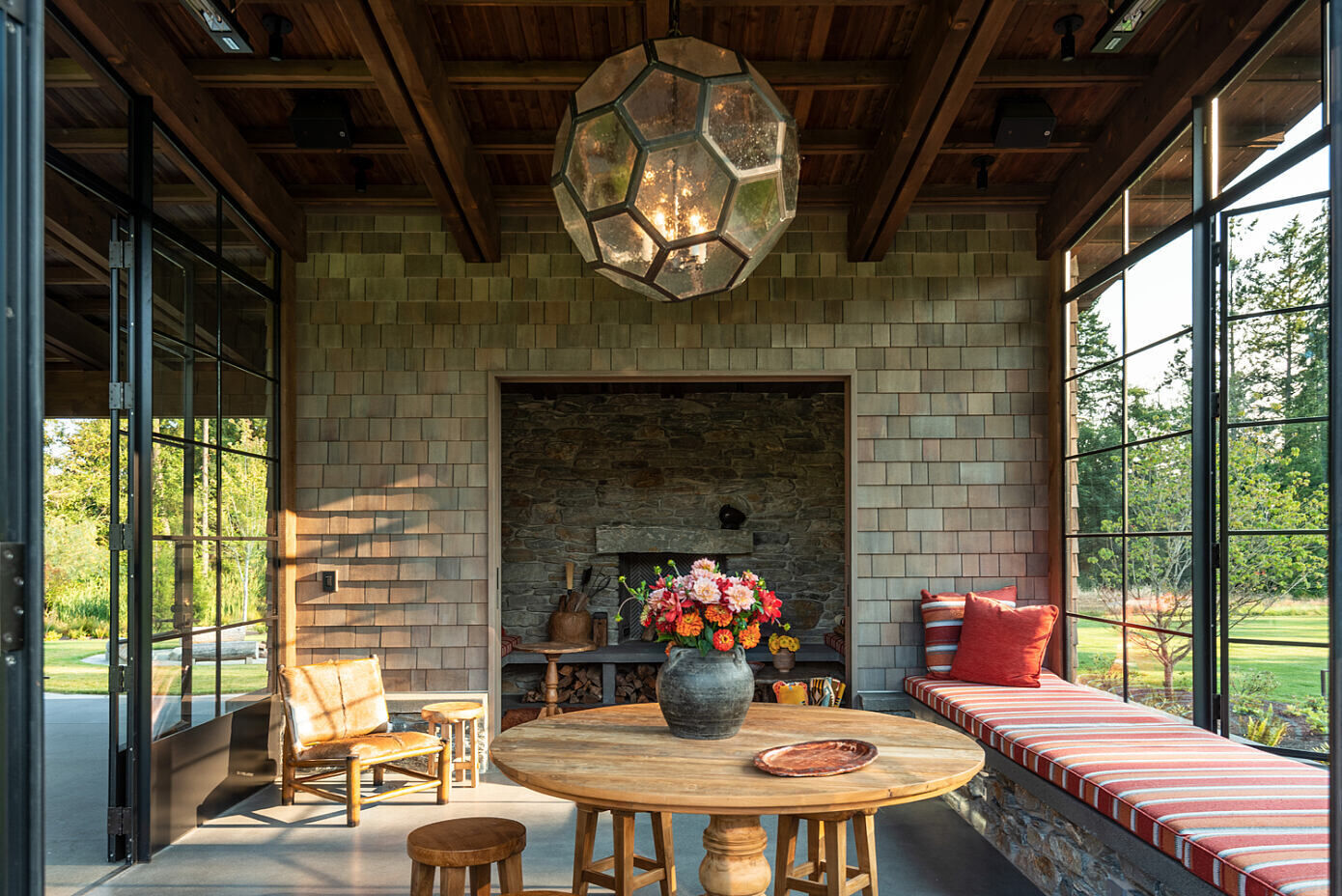 A light filled sunroom with wrap around seating flows into a cozy inglenook fireplace, creating intimate gathering spaces within the larger pavilion.