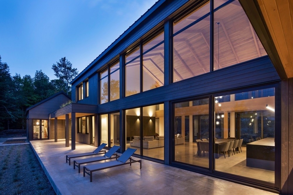 A spacious open plan great room features long walls of windows, offering forest views on one side and lake views on the other.