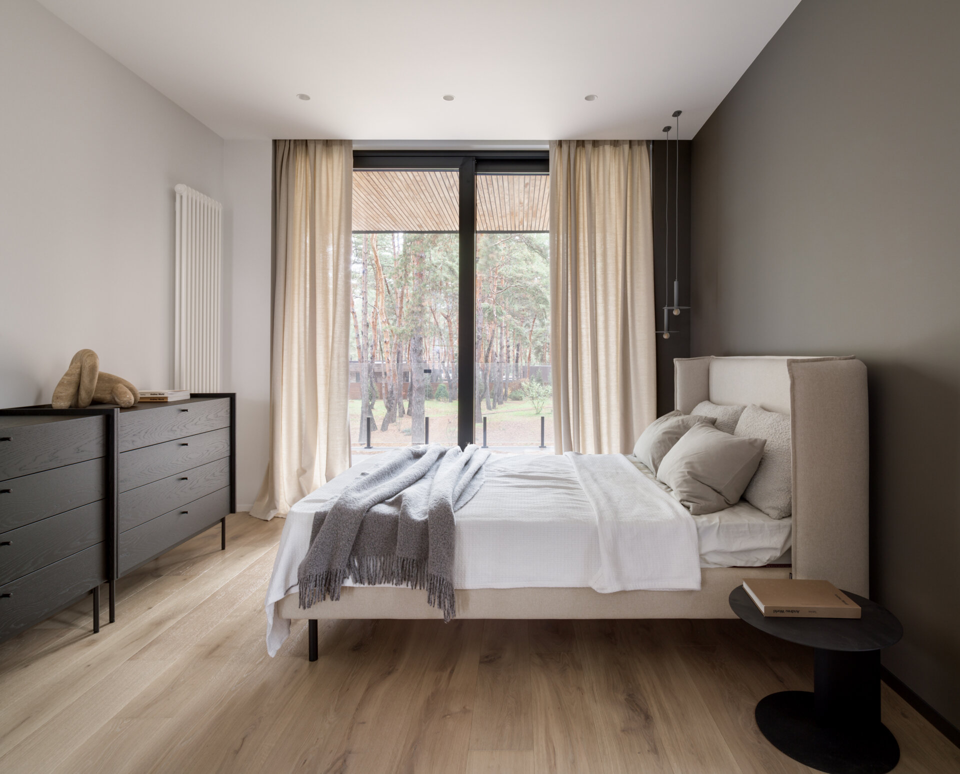 This modern bedroom is composed as a quiet space, defined by soft beige upholstery and light wood floors. Sheer curtains soften daylight from the wide glass doors and frame views of the surrounding trees.