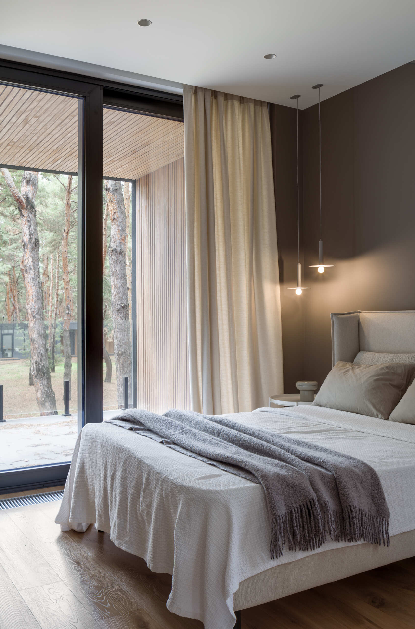 This modern bedroom is composed as a quiet space, defined by soft beige upholstery and light wood floors. Sheer curtains soften daylight from the wide glass doors and frame views of the surrounding trees.