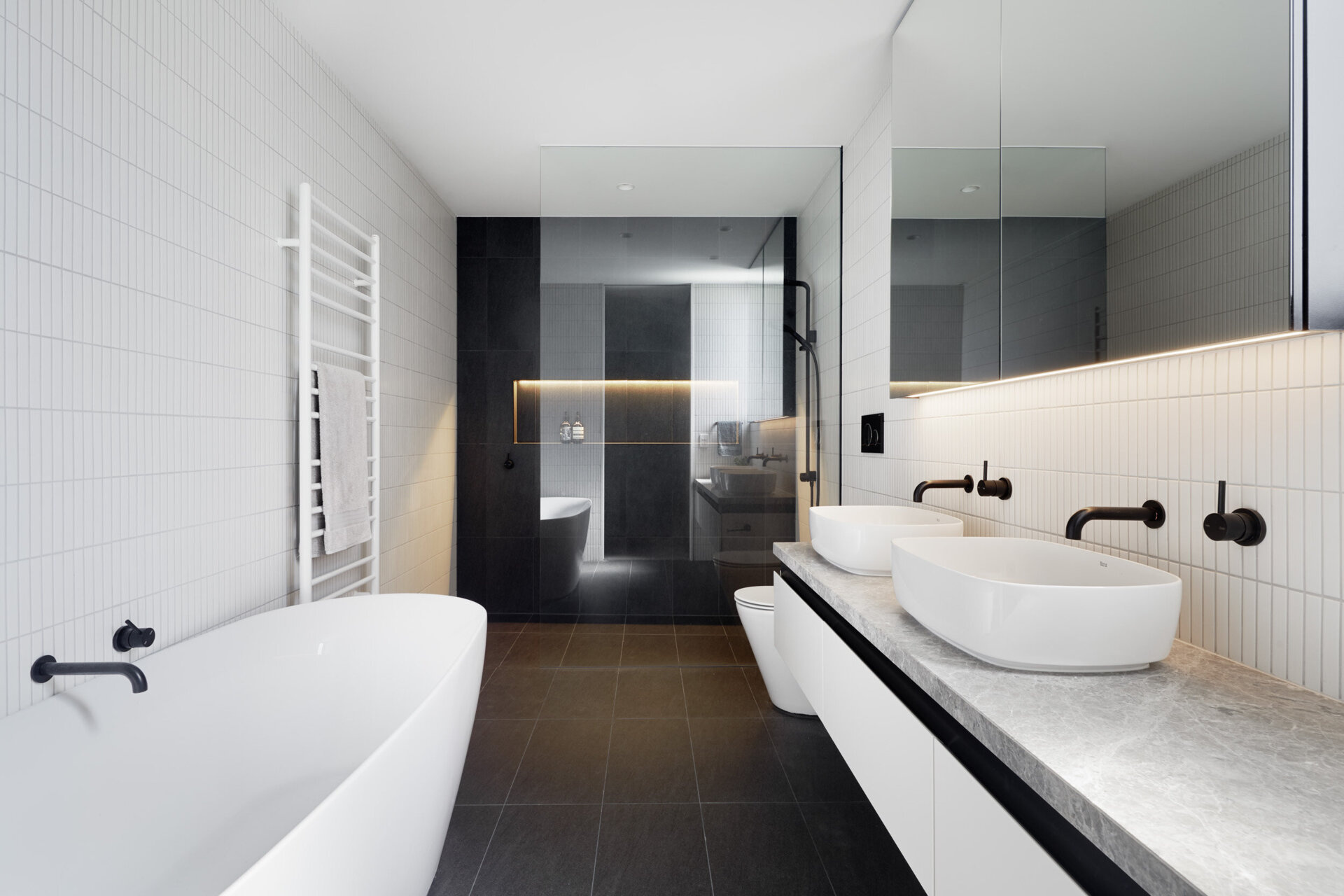 Black shower tiles paired with hidden niche lighting create a bold focal point, contrasted by crisp white finishes throughout the rest of the bathroom.