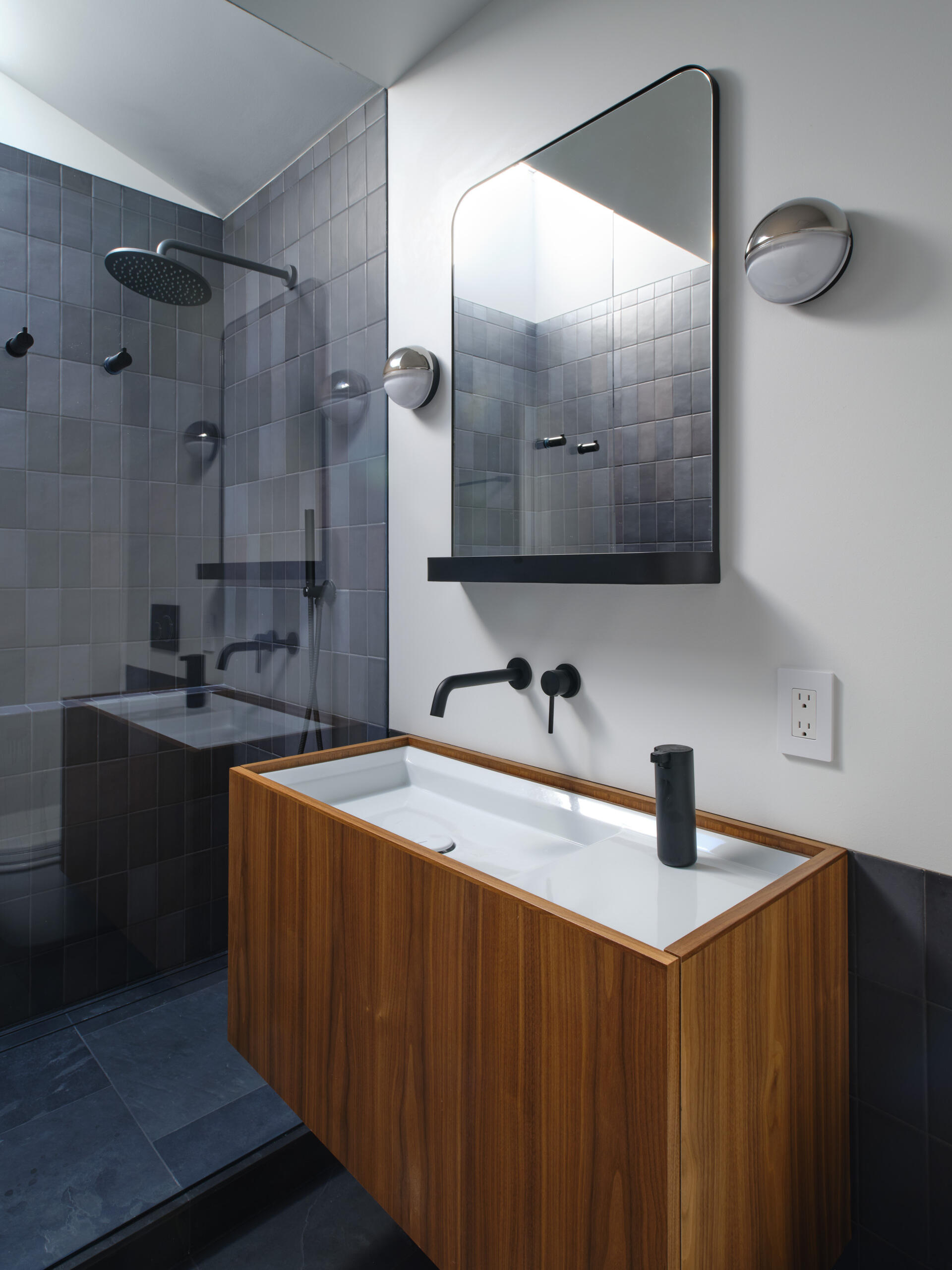 Like many older homes, the bathrooms were previously dark and enclosed. Natural light was minimal. The reconfigured main bathroom introduces a dramatic skylight directly over the shower. Dark gray tiles, slate flooring, and walnut cabinetry create contrast beneath the daylight above.