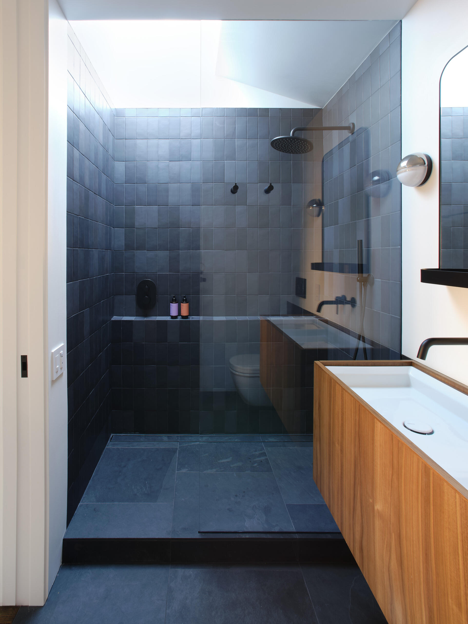 Like many older homes, the bathrooms were previously dark and enclosed. Natural light was minimal. The reconfigured main bathroom introduces a dramatic skylight directly over the shower. Dark gray tiles, slate flooring, and walnut cabinetry create contrast beneath the daylight above.