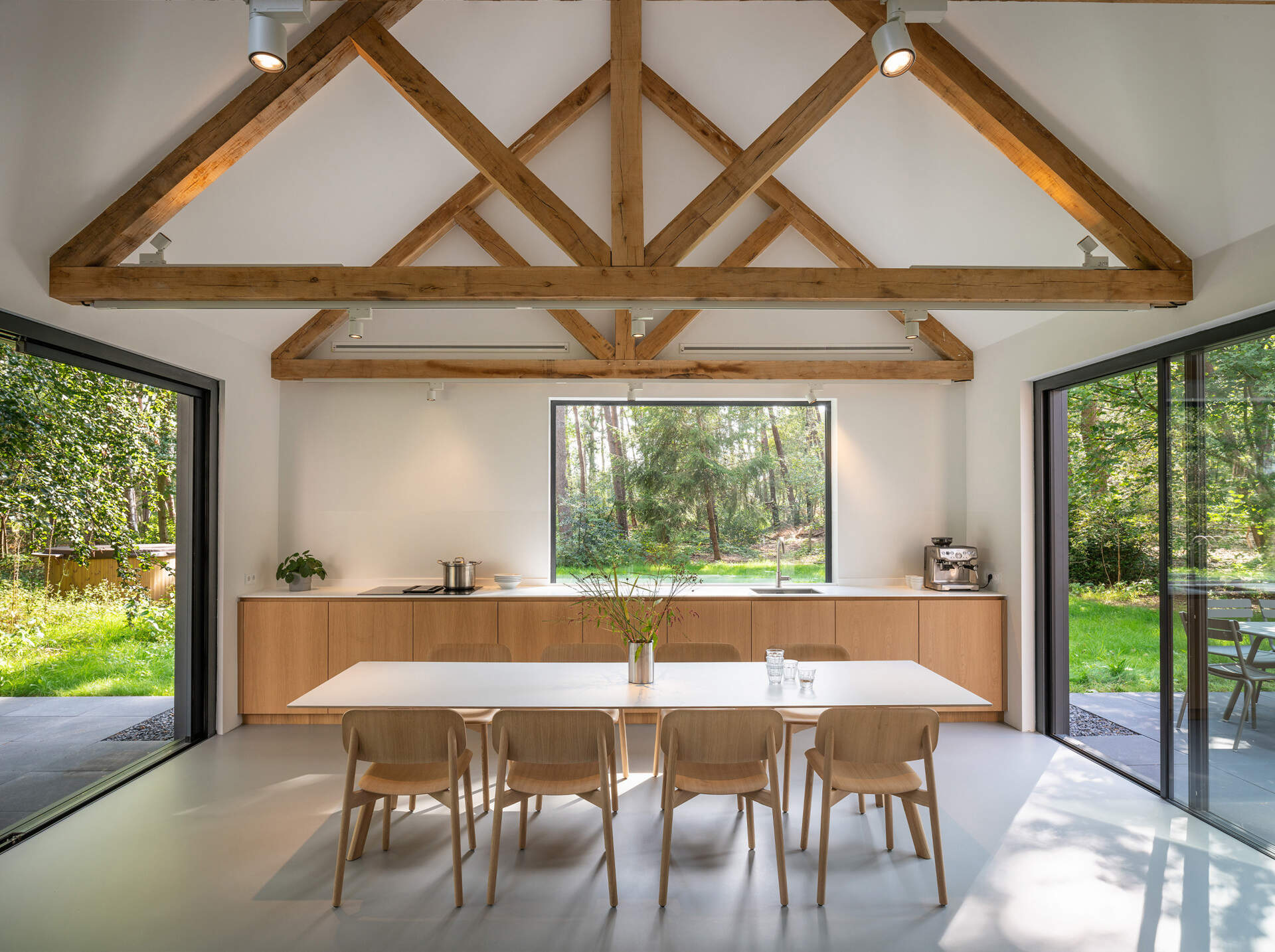 This minimalist kitchen, dining, and living area shares one light-filled space with exposed timber beams and black-framed sliding glass doors that open directly to the surrounding woods.
