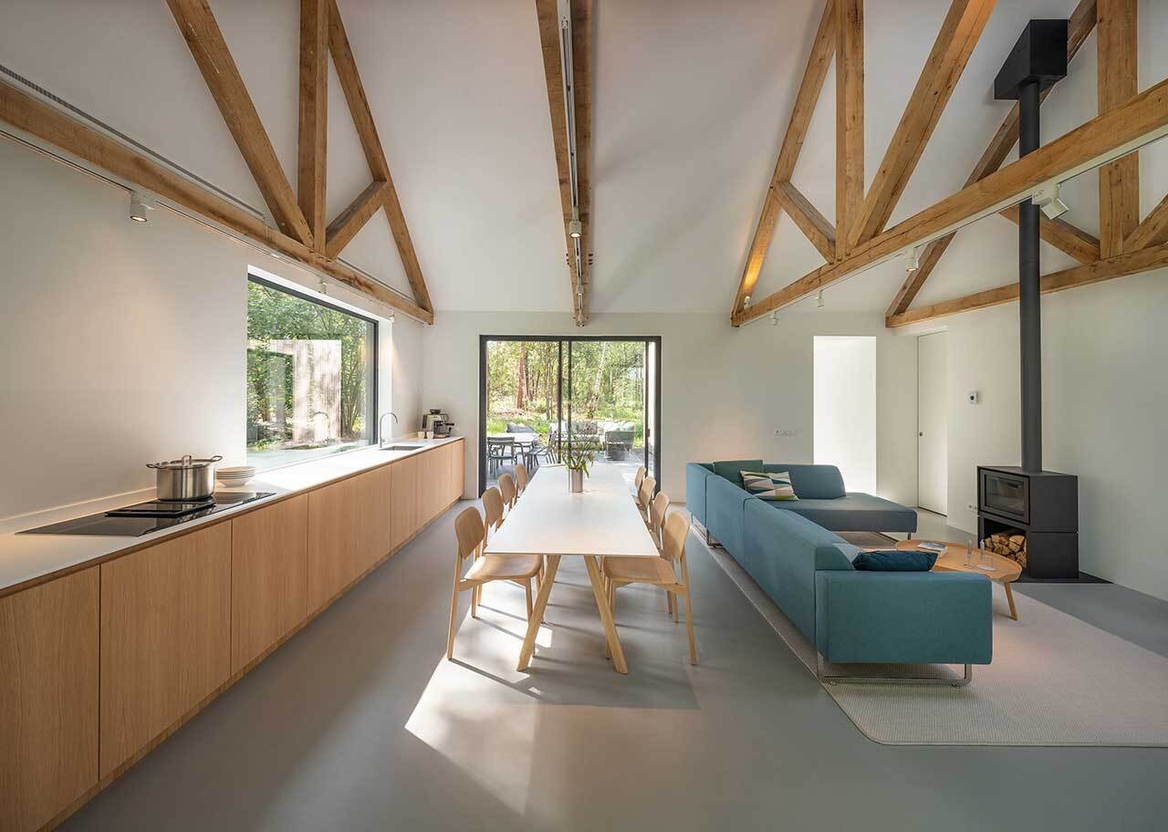 This minimalist kitchen, dining, and living area shares one light-filled space with exposed timber beams and black-framed sliding glass doors that open directly to the surrounding woods.