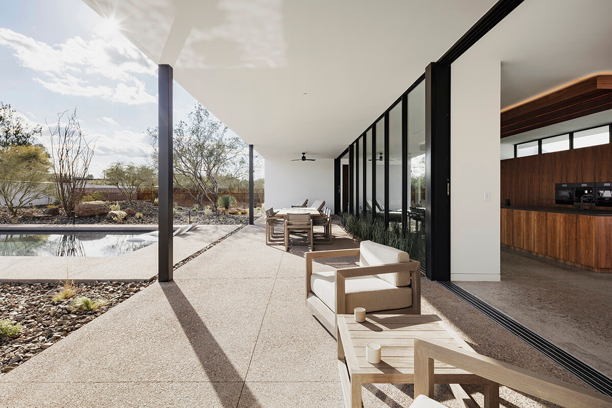 A covered patio shifts from shaded daytime refuge to softly lit evening lounge. The lap pool extends beyond the home’s “O” shape and ends in a built-in fire pit bench for gatherings.
