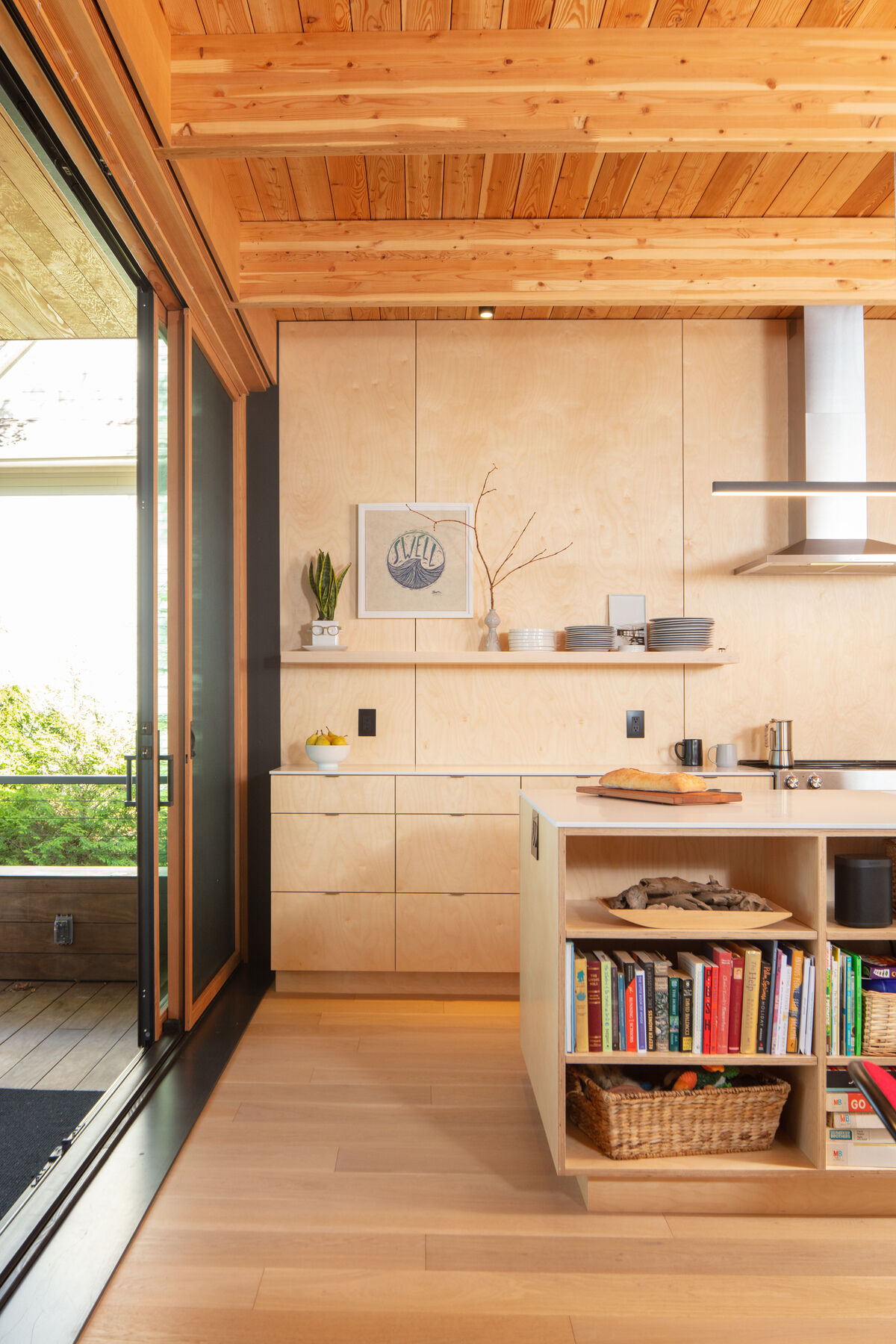 This airy kitchen leans into a soft, natural palette, with pale wood cabinetry and wide-plank floors that keep everything feeling light and easy. The standout ceiling, lined in warm wood boards and beams, adds texture overhead, especially with the simple black spotlights.