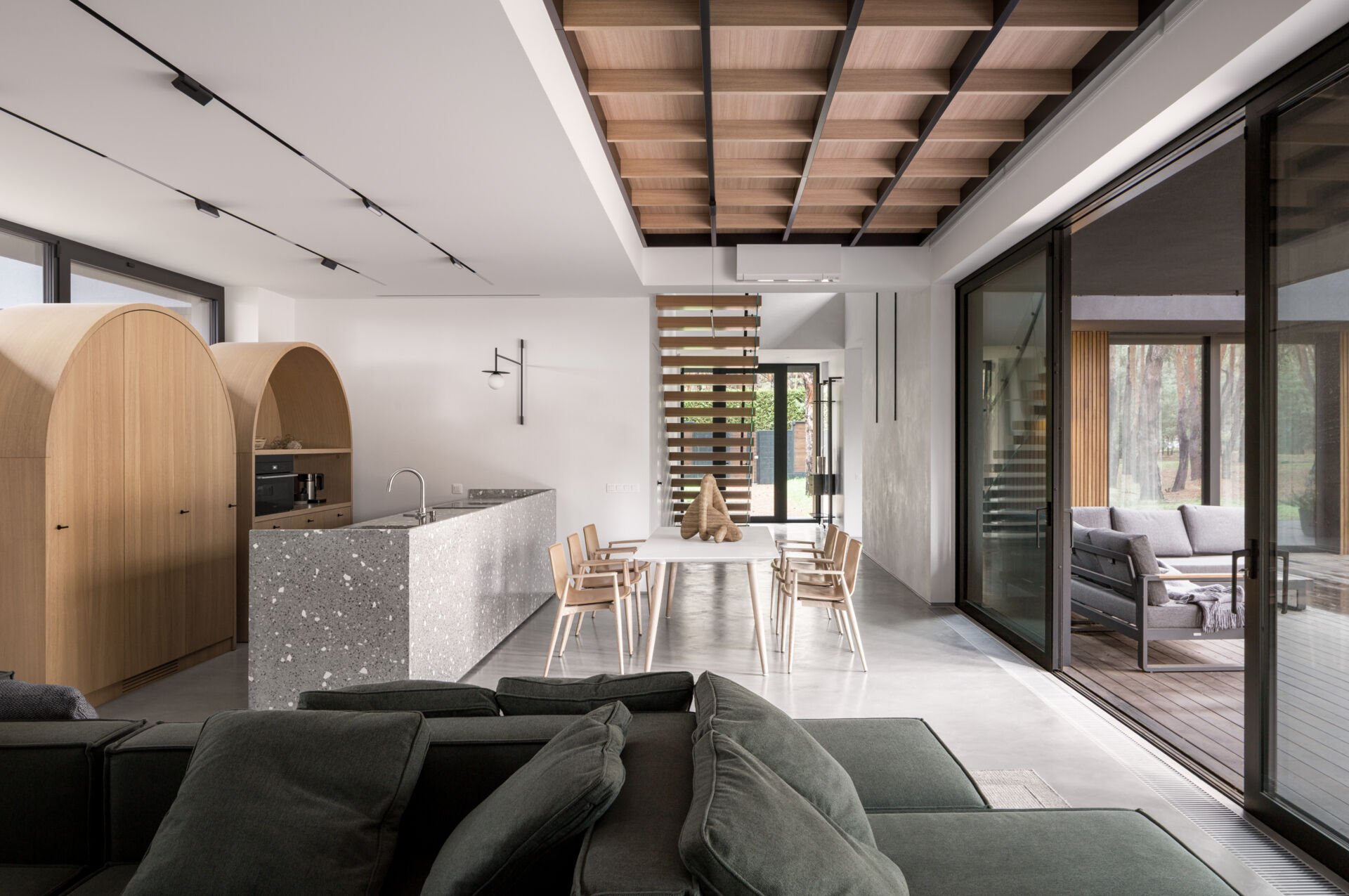 This open-plan kitchen and dining space features smooth concrete floors, bright white walls, and warm wood details that keep it inviting. The speckled terrazzo island adds texture and a touch of playfulness, echoed by the arched wood cabinets that soften the modern lines, while a simple white table and light wood chairs define the dining area.