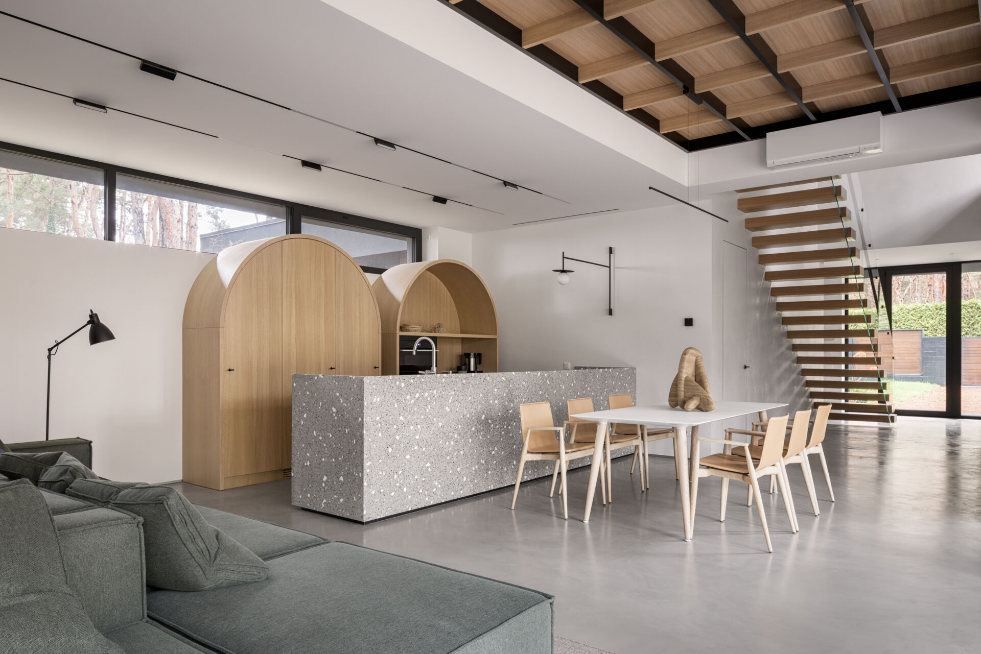 This open-plan kitchen and dining space features smooth concrete floors, bright white walls, and warm wood details that keep it inviting. The speckled terrazzo island adds texture and a touch of playfulness, echoed by the arched wood cabinets that soften the modern lines, while a simple white table and light wood chairs define the dining area.