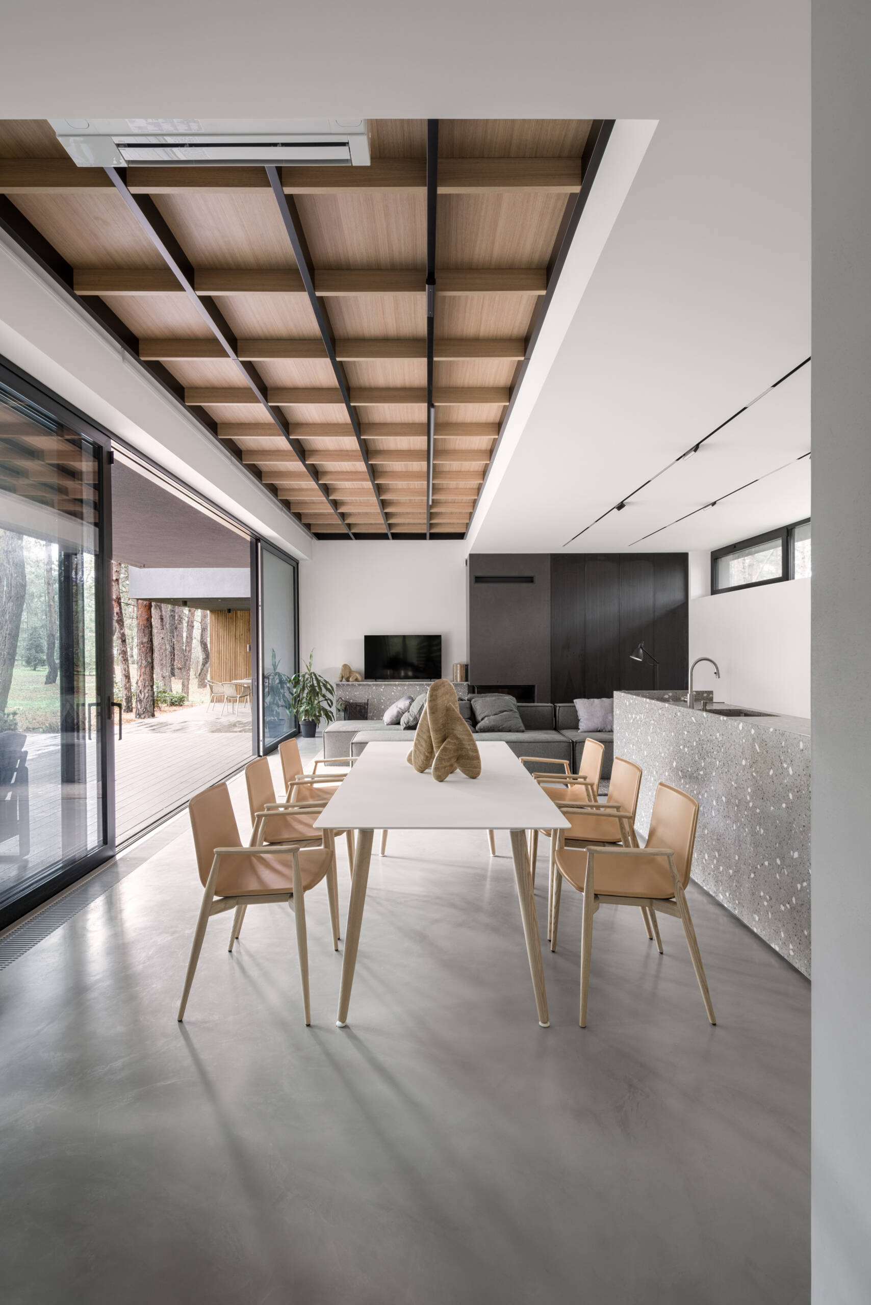 This open-plan kitchen and dining space features smooth concrete floors, bright white walls, and warm wood details that keep it inviting. The speckled terrazzo island adds texture and a touch of playfulness, echoed by the arched wood cabinets that soften the modern lines, while a simple white table and light wood chairs define the dining area.