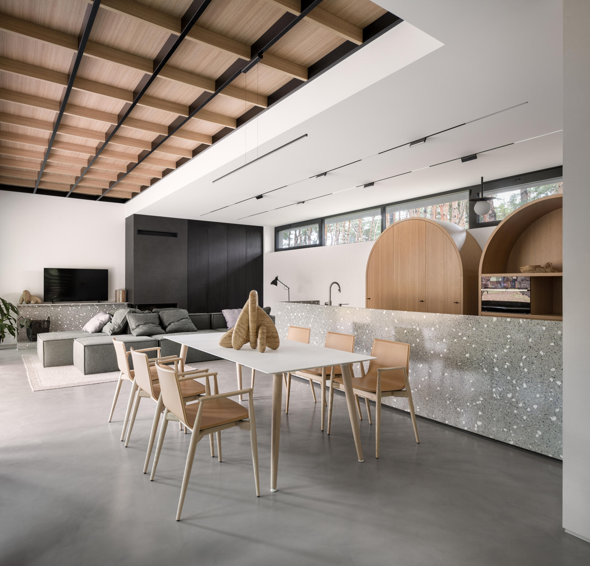 This open-plan kitchen and dining space features smooth concrete floors, bright white walls, and warm wood details that keep it inviting. The speckled terrazzo island adds texture and a touch of playfulness, echoed by the arched wood cabinets that soften the modern lines, while a simple white table and light wood chairs define the dining area.