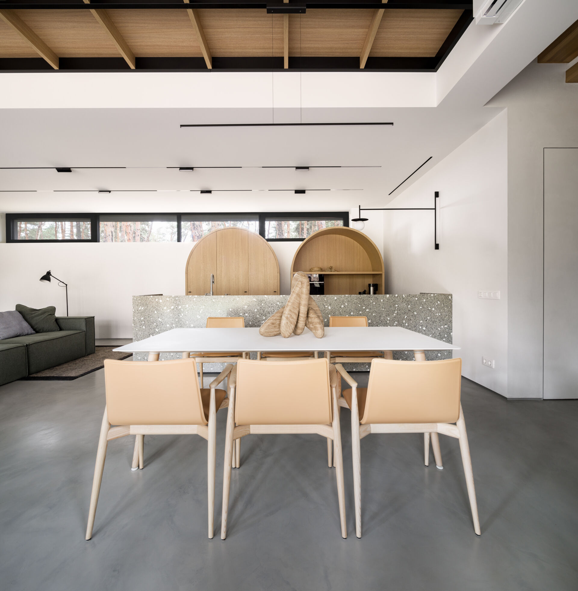 This open-plan kitchen and dining space features smooth concrete floors, bright white walls, and warm wood details that keep it inviting. The speckled terrazzo island adds texture and a touch of playfulness, echoed by the arched wood cabinets that soften the modern lines, while a simple white table and light wood chairs define the dining area.
