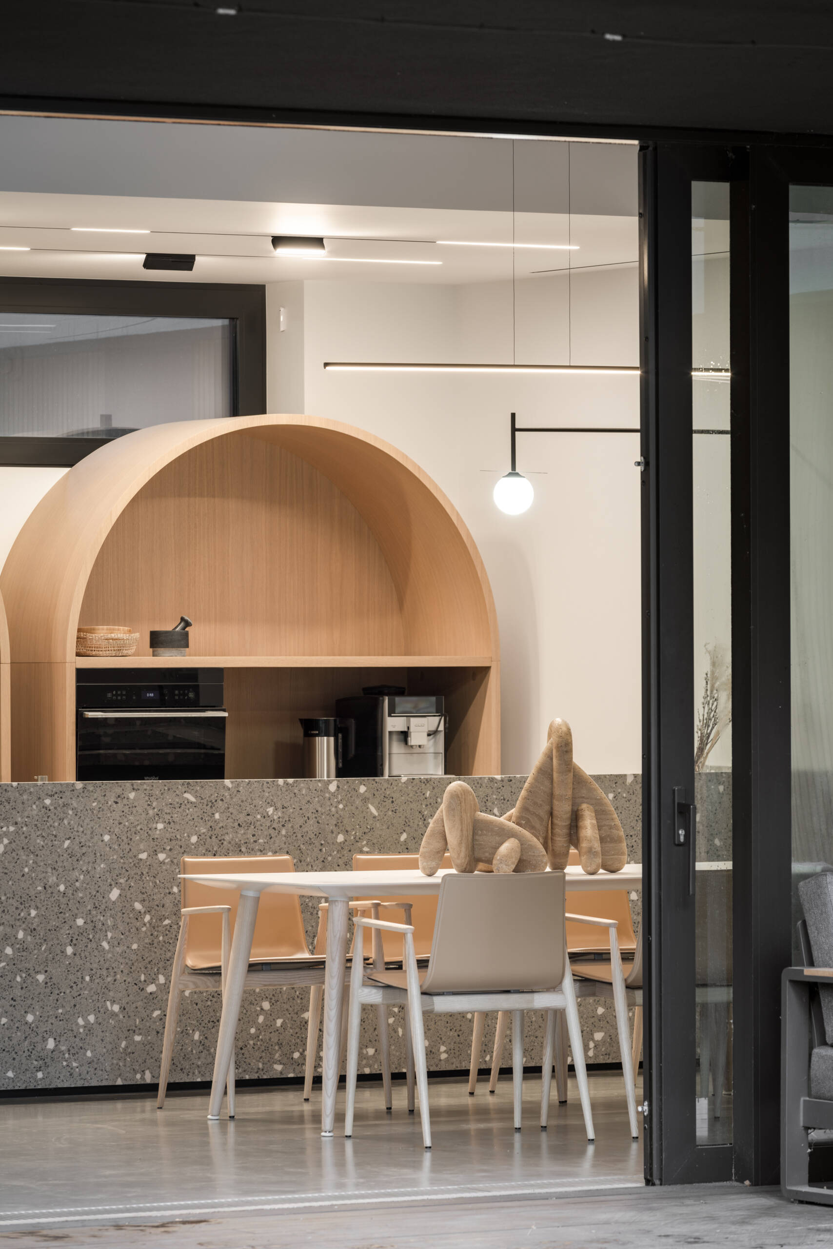 This open-plan kitchen and dining space features smooth concrete floors, bright white walls, and warm wood details that keep it inviting. The speckled terrazzo island adds texture and a touch of playfulness, echoed by the arched wood cabinets that soften the modern lines, while a simple white table and light wood chairs define the dining area.