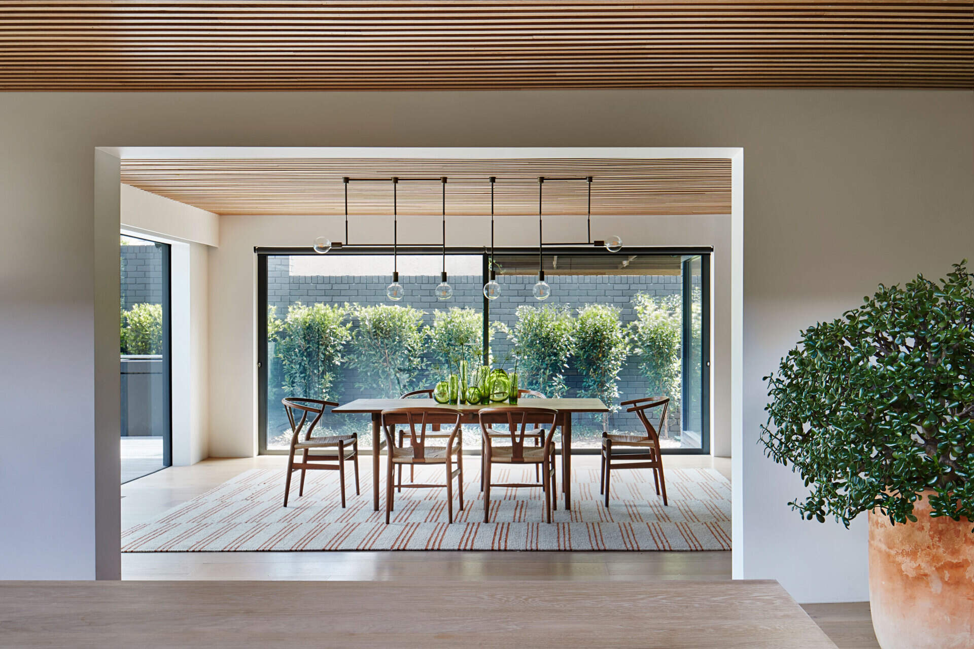 Plate glass walls bring garden views into the dining space, while minimalist lighting adds quiet structure above the table.