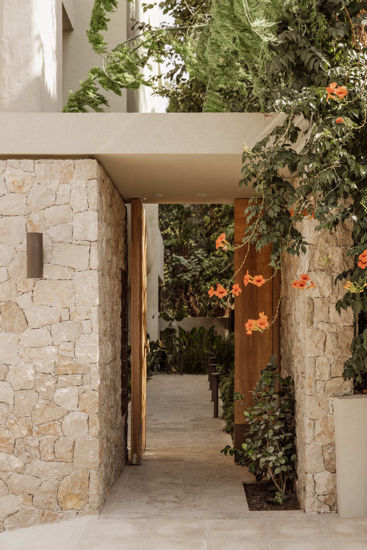 The approach to the building entrance sets the tone immediately. A timber gate opens onto a narrow, planted pathway where soft greenery lines a pale stone walkway, guiding visitors forward at an unhurried pace. Overhead, the building’s curved form gently overhangs the path, casting shifting shadows across textured walls.