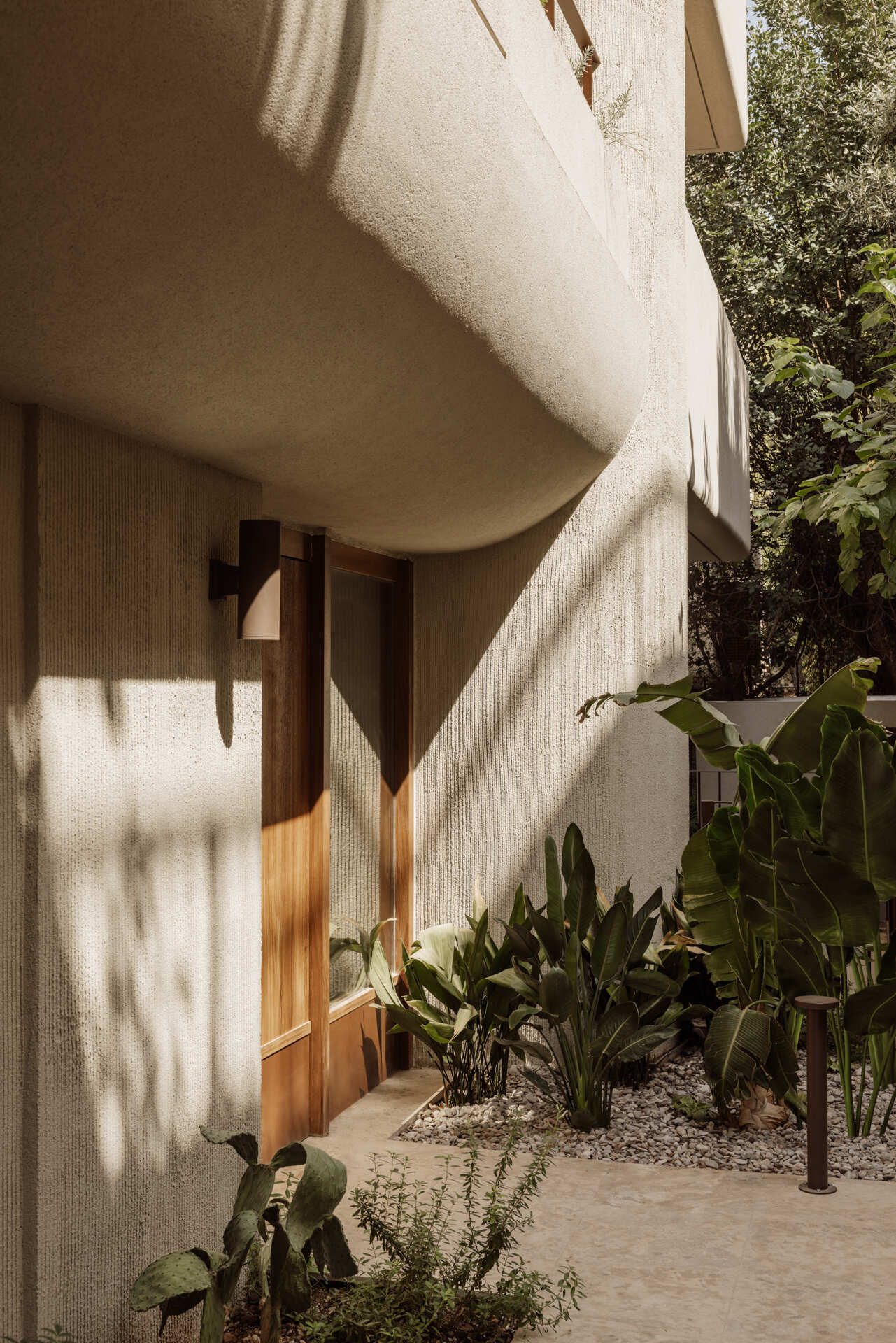 The approach to the building entrance sets the tone immediately. A timber gate opens onto a narrow, planted pathway where soft greenery lines a pale stone walkway, guiding visitors forward at an unhurried pace. Overhead, the building’s curved form gently overhangs the path, casting shifting shadows across textured walls.