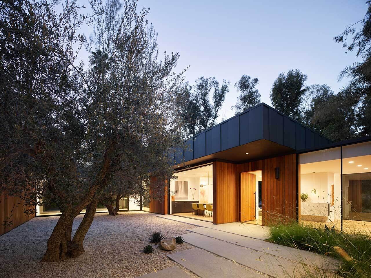 Una casa baja en Los Ángeles está revestida con cedro rojo occidental, tablas y listones de carbón, con adoquines de concreto y pasto silvestre que conducen a un patio de olivos.