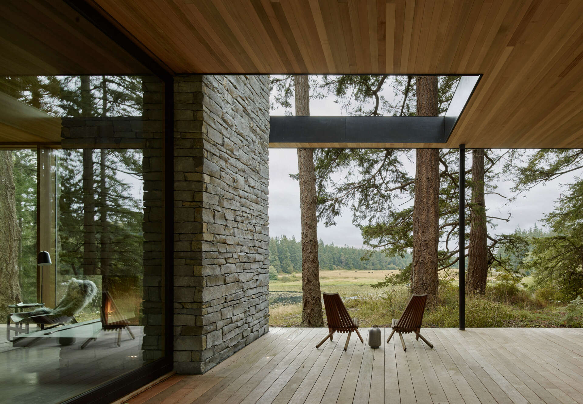A stone fireplace with a concrete hearth anchors this open plan living room, where sliding glass walls connect the interior to a covered deck and sweeping countryside views.
