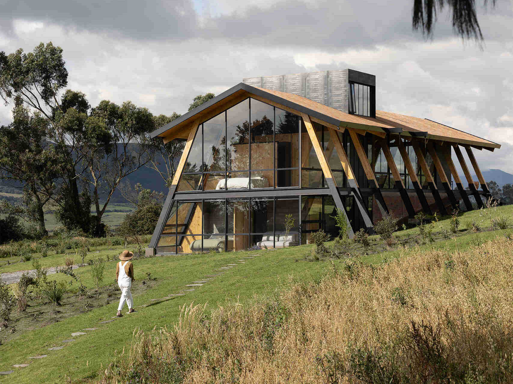 Ten wooden ribs define the rhythm of this Ecuadorian home. Large eaves, a brick base, and a covered ramp shape how the house meets the slope and protects against sun and rain.