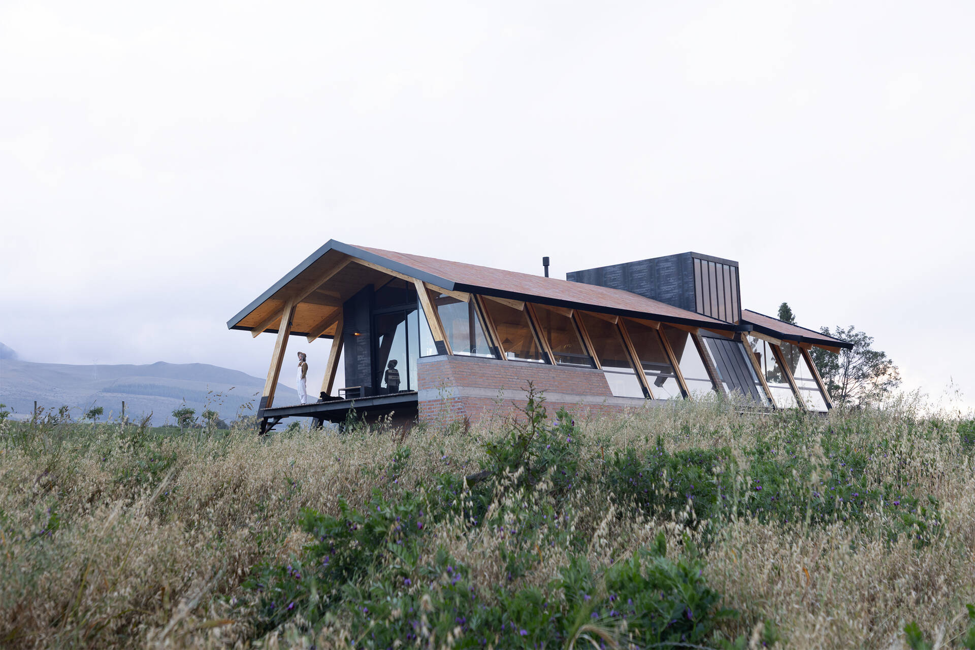 Ten wooden ribs define the rhythm of this Ecuadorian home. Large eaves, a brick base, and a covered ramp shape how the house meets the slope and protects against sun and rain.