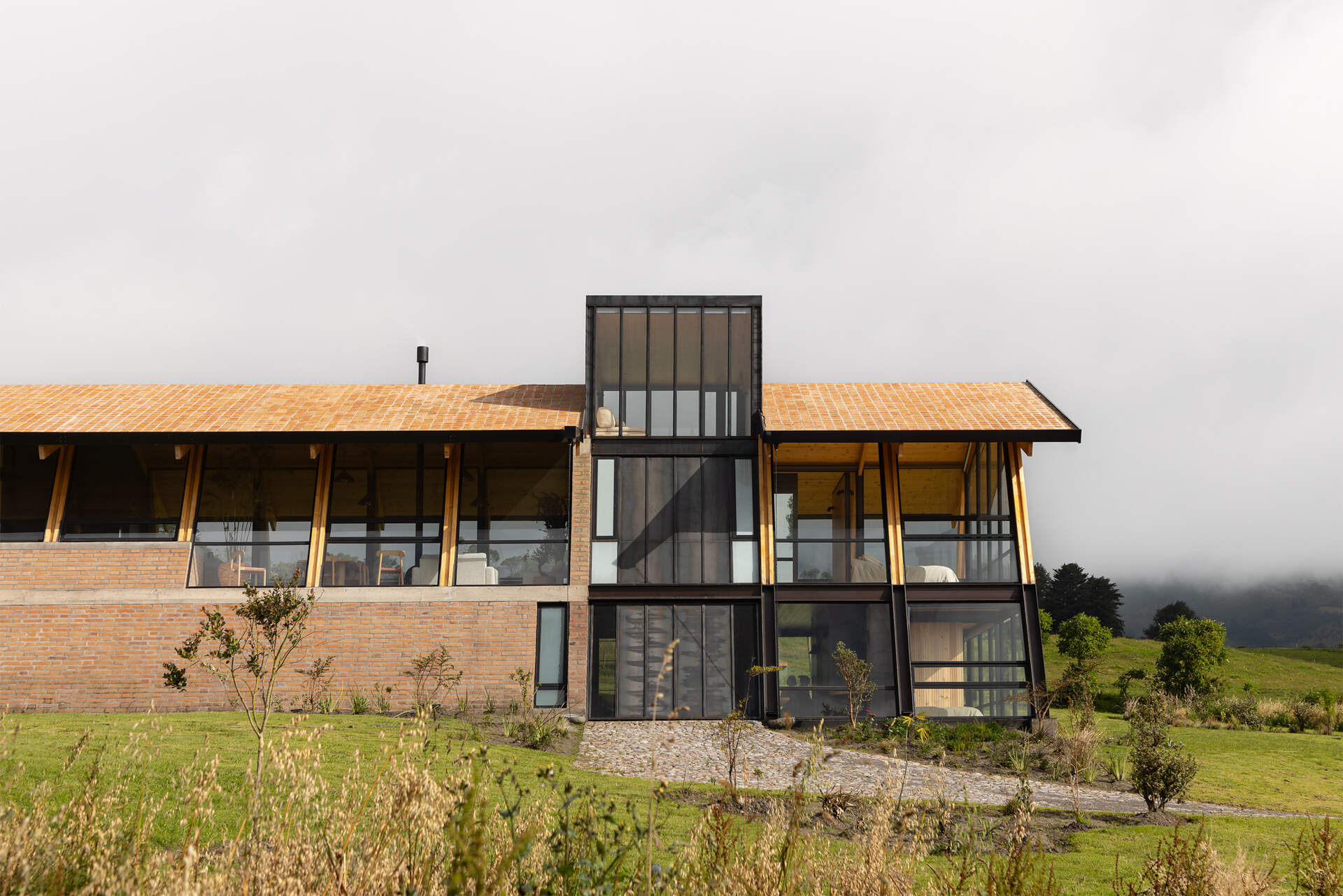 Ten wooden ribs define the rhythm of this Ecuadorian home. Large eaves, a brick base, and a covered ramp shape how the house meets the slope and protects against sun and rain.