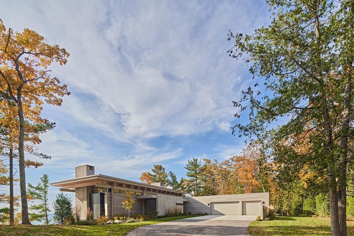 The entryway is hidden behind a concrete wall, guiding visitors through the four-season room to stunning lake views. The driveway and garage maintain privacy while creating a welcoming approach.