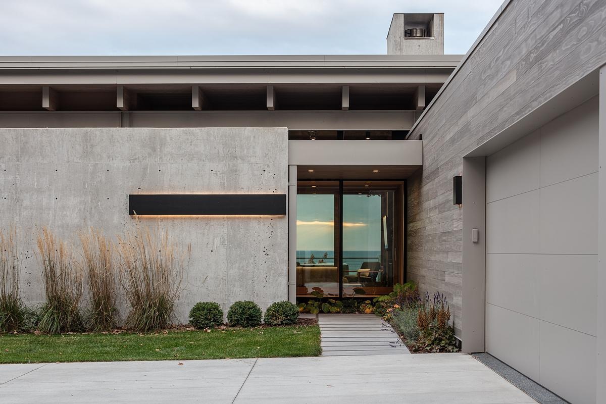 The entryway is hidden behind a concrete wall, guiding visitors through the four-season room to stunning lake views. The driveway and garage maintain privacy while creating a welcoming approach.