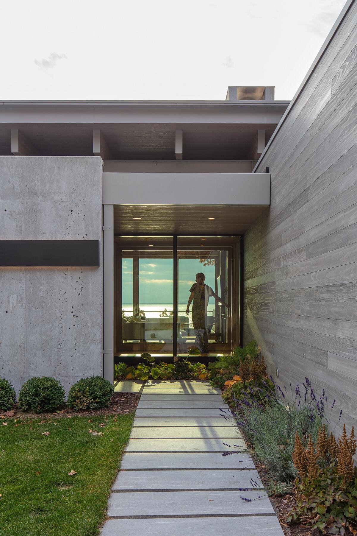 The entryway is hidden behind a concrete wall, guiding visitors through the four-season room to stunning lake views. The driveway and garage maintain privacy while creating a welcoming approach.
