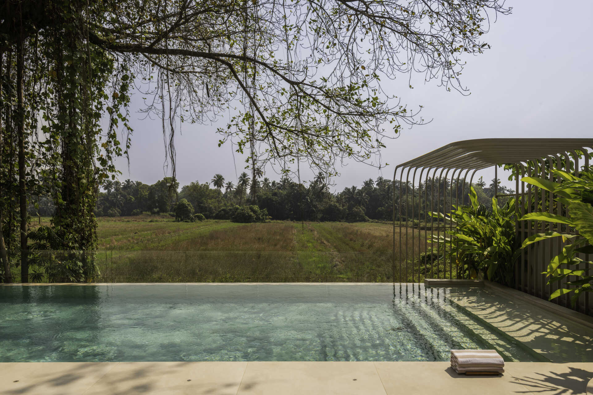 A stepped water channel leads to a reflective pool overlooking open fields in Goa. Seamless indoor-outdoor flooring and an in-situ concrete jacuzzi create a calm, continuous connection between architecture and landscape.