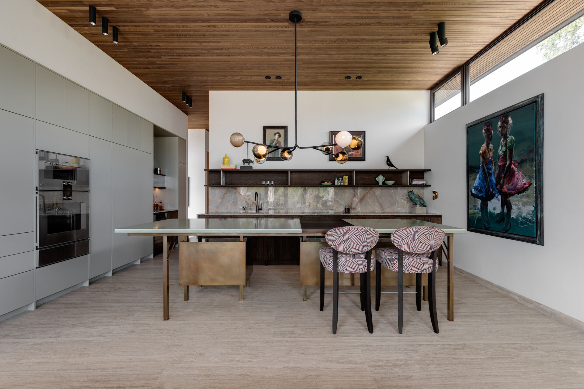 In the kitchen, minimalist grey cabinets line the wall, while an island with copper accents is positioned beneath a chandelier and nearby artwork adds color to the space.