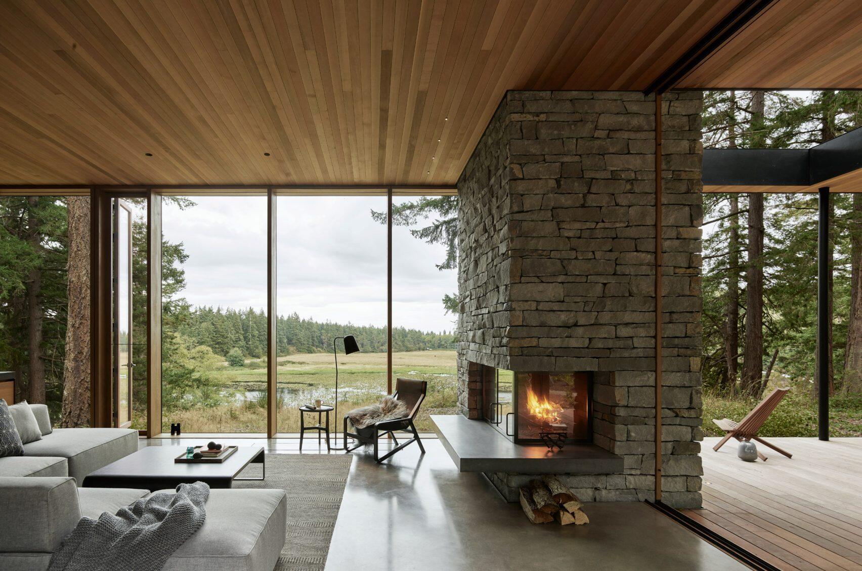A stone fireplace with a concrete hearth anchors this open plan living room, where sliding glass walls connect the interior to a covered deck and sweeping countryside views.