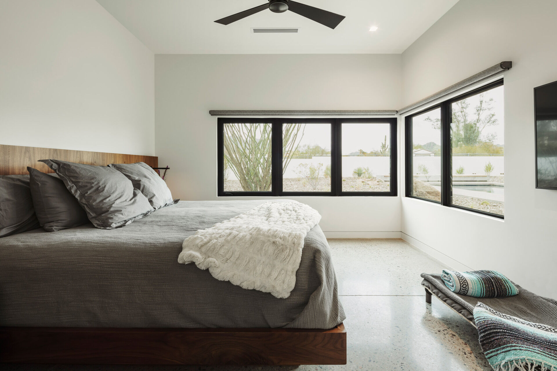 The main bedroom continues the language of integration. A built-in wood bed frame and headboard anchor the space, while windows on each wall look toward the pool, keeping water and sky in constant view.