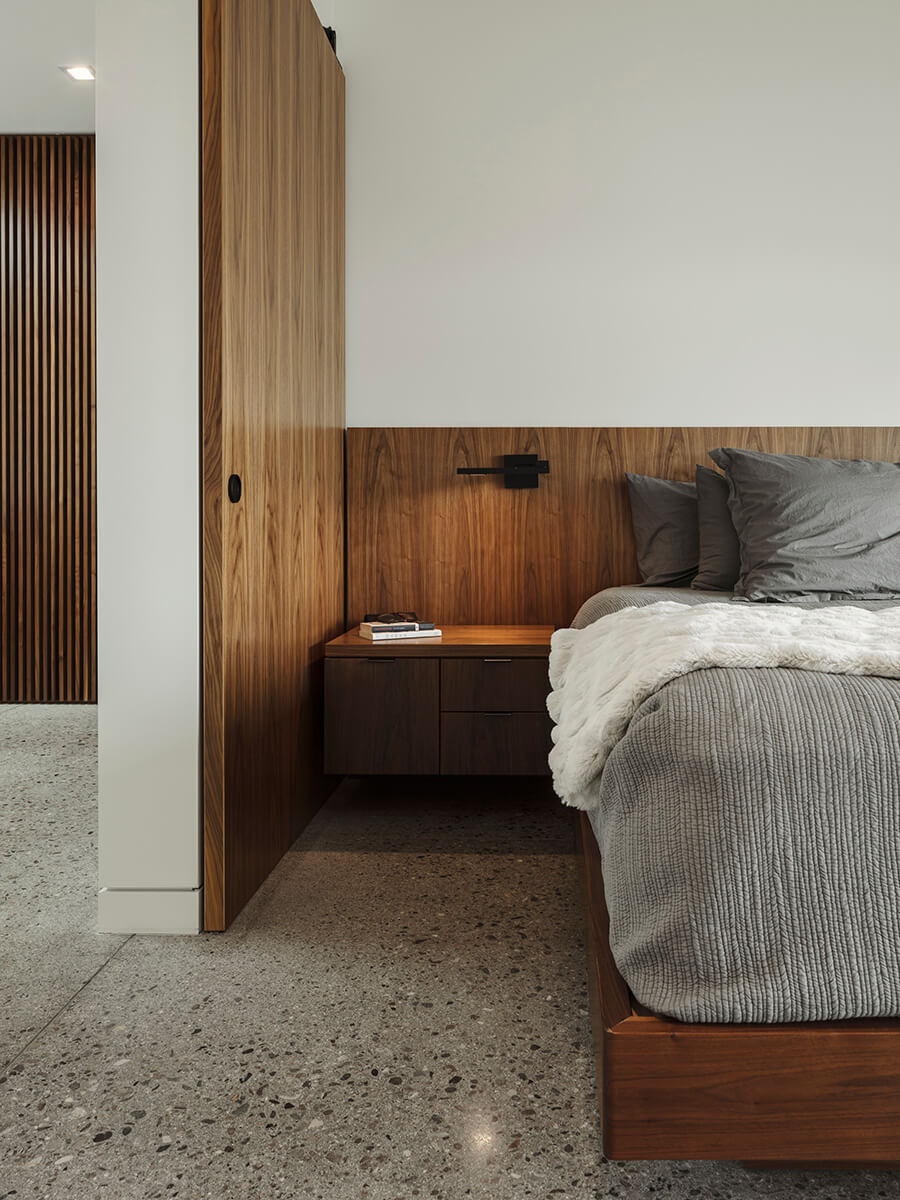 The main bedroom continues the language of integration. A built-in wood bed frame and headboard anchor the space, while windows on each wall look toward the pool, keeping water and sky in constant view.