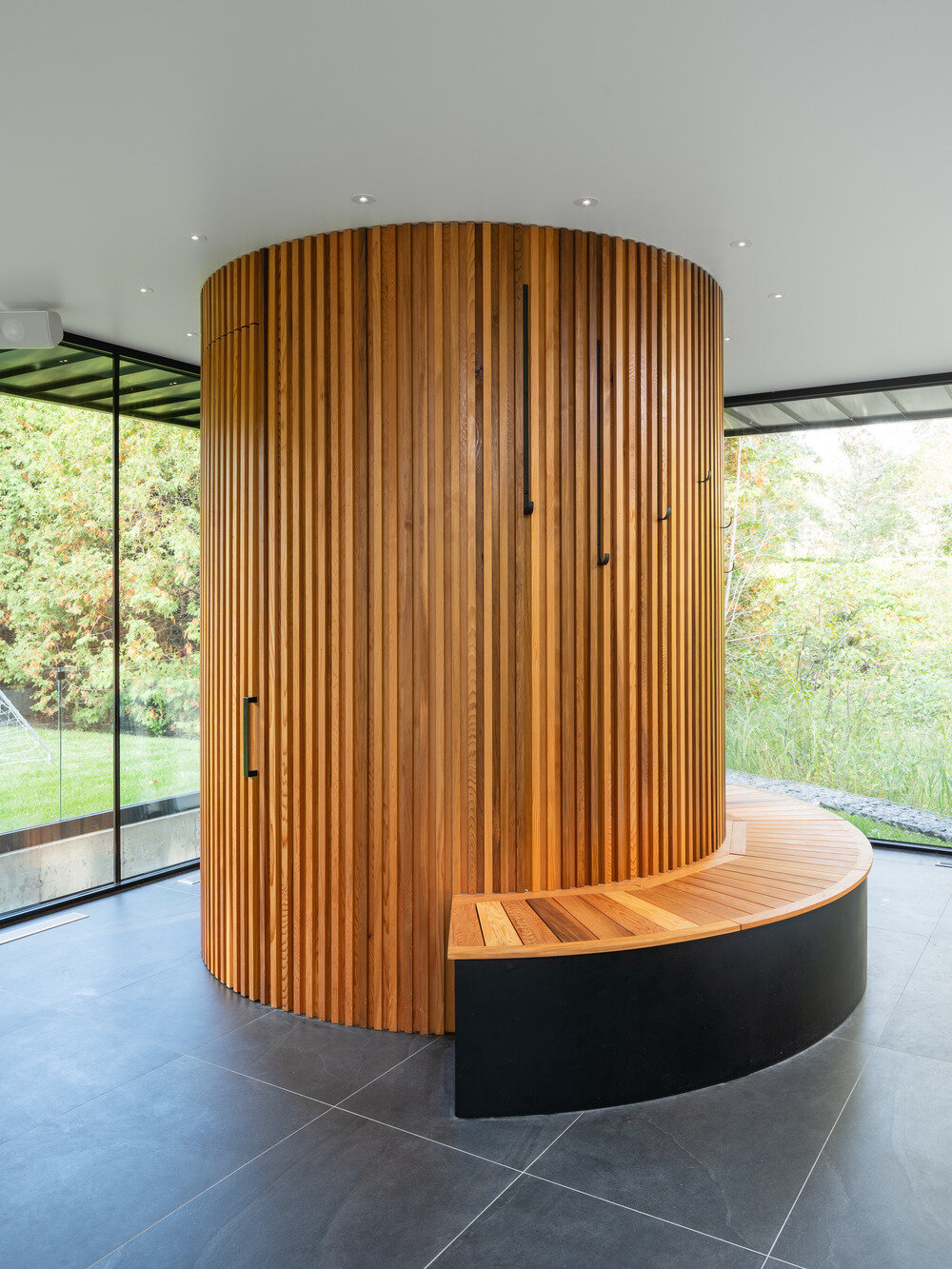 A cylindrical bathroom clad in red cedar stands as a warm sculptural feature within the glass pool house, complete with a curved bench and hidden entrance.