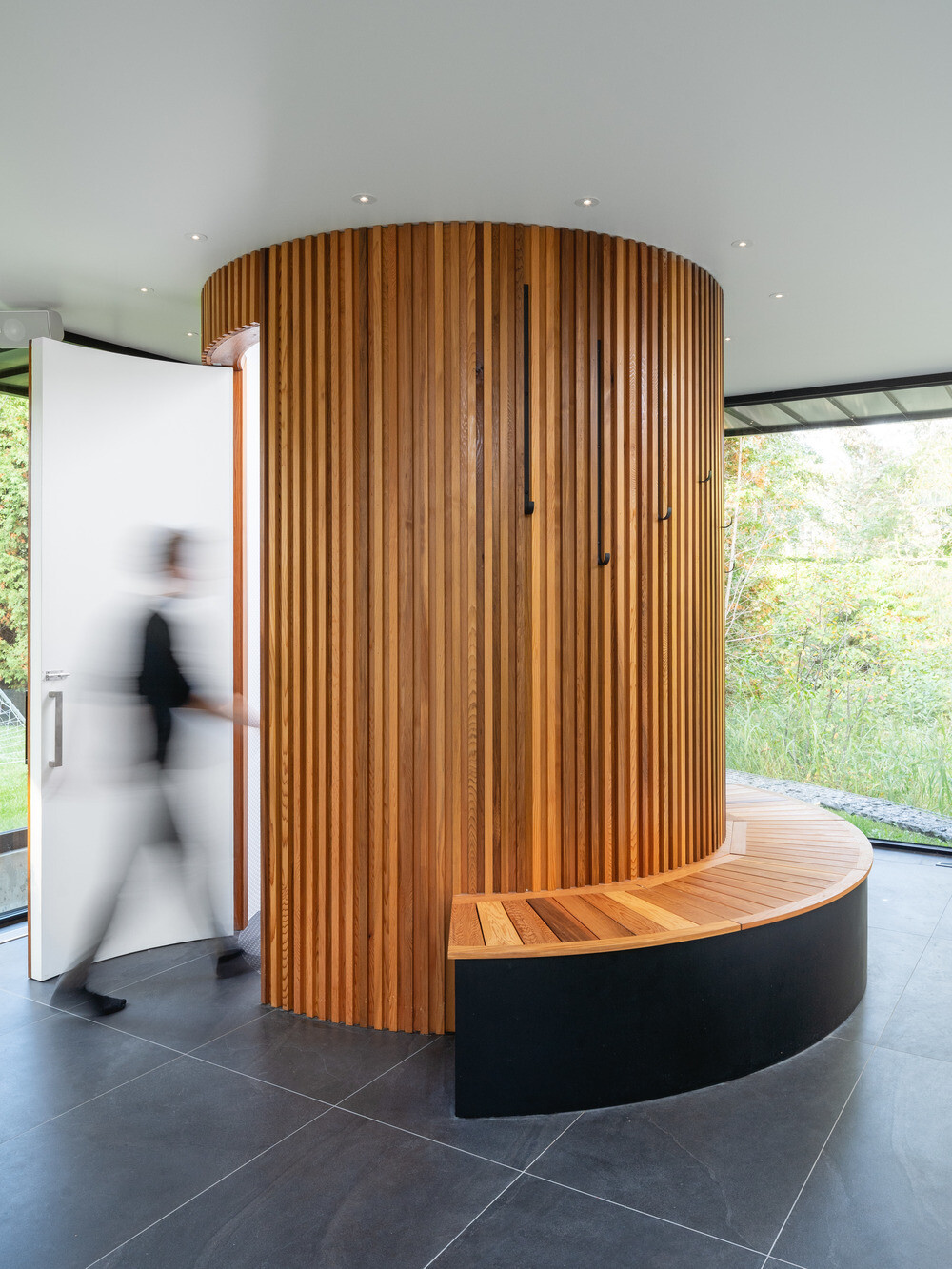 A cylindrical bathroom clad in red cedar stands as a warm sculptural feature within the glass pool house, complete with a curved bench and hidden entrance.