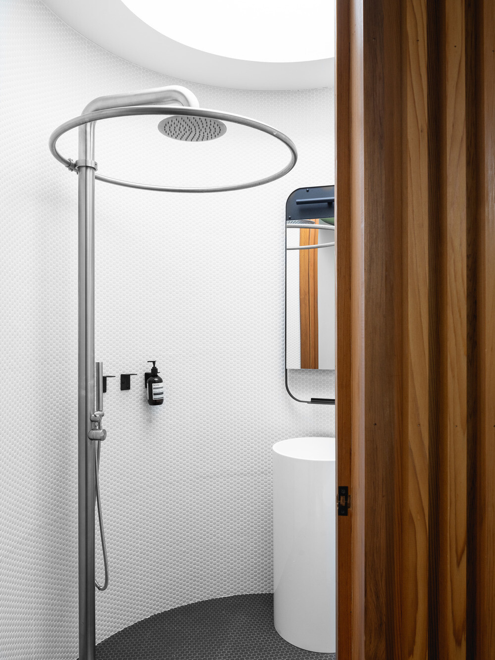 A cylindrical bathroom clad in red cedar stands as a warm sculptural feature within the glass pool house, complete with a curved bench and hidden entrance.