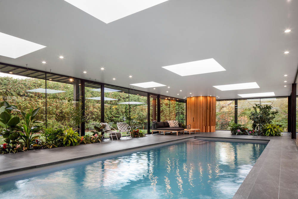Skylights flood the interior with natural light while a lounging deck and layers of tropical plants create an oasis feel inside this modern glass pool house.