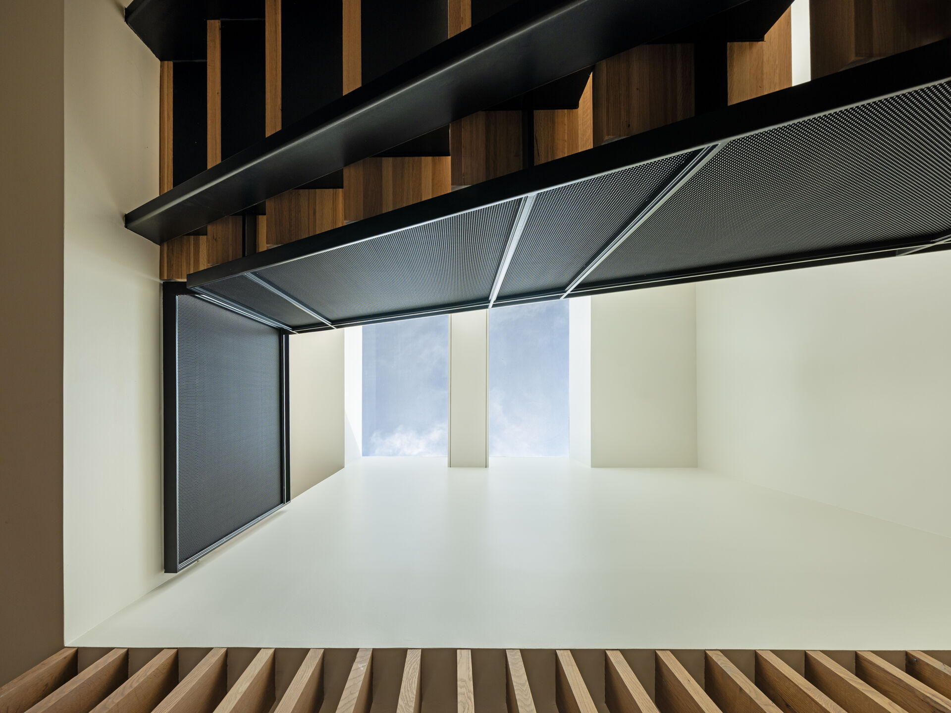 A vertical wood batten screen filters light from a central skylight, animating the stair core with soft, shifting shadows. The staircase connects all levels while continuing the exterior material palette indoors.