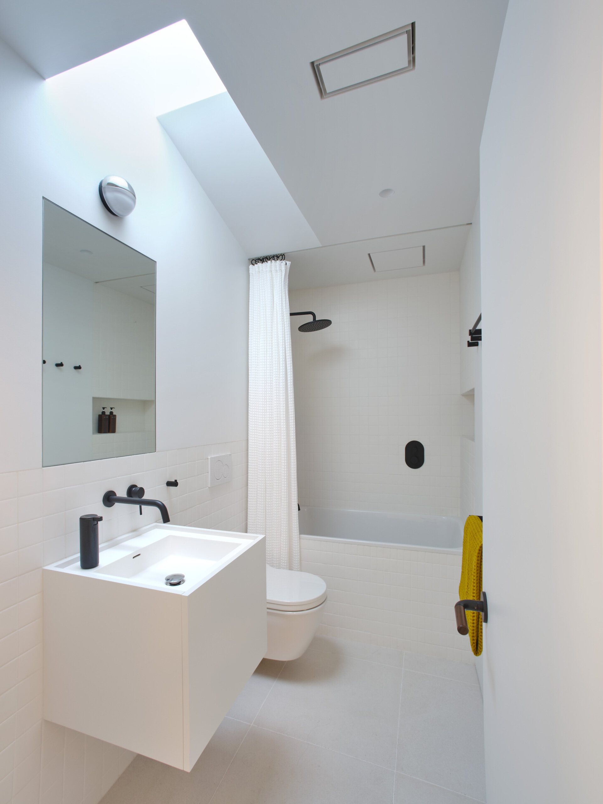 Before renovation, the guest bathroom shared the same limitations of low light and constrained proportions. In response, the design takes an opposite tonal approach to the main bath. Varied sizes of white stack bond tiles create graphic interest while visually enlarging the modest room. The brighter palette amplifies available light, making the space feel more expansive.