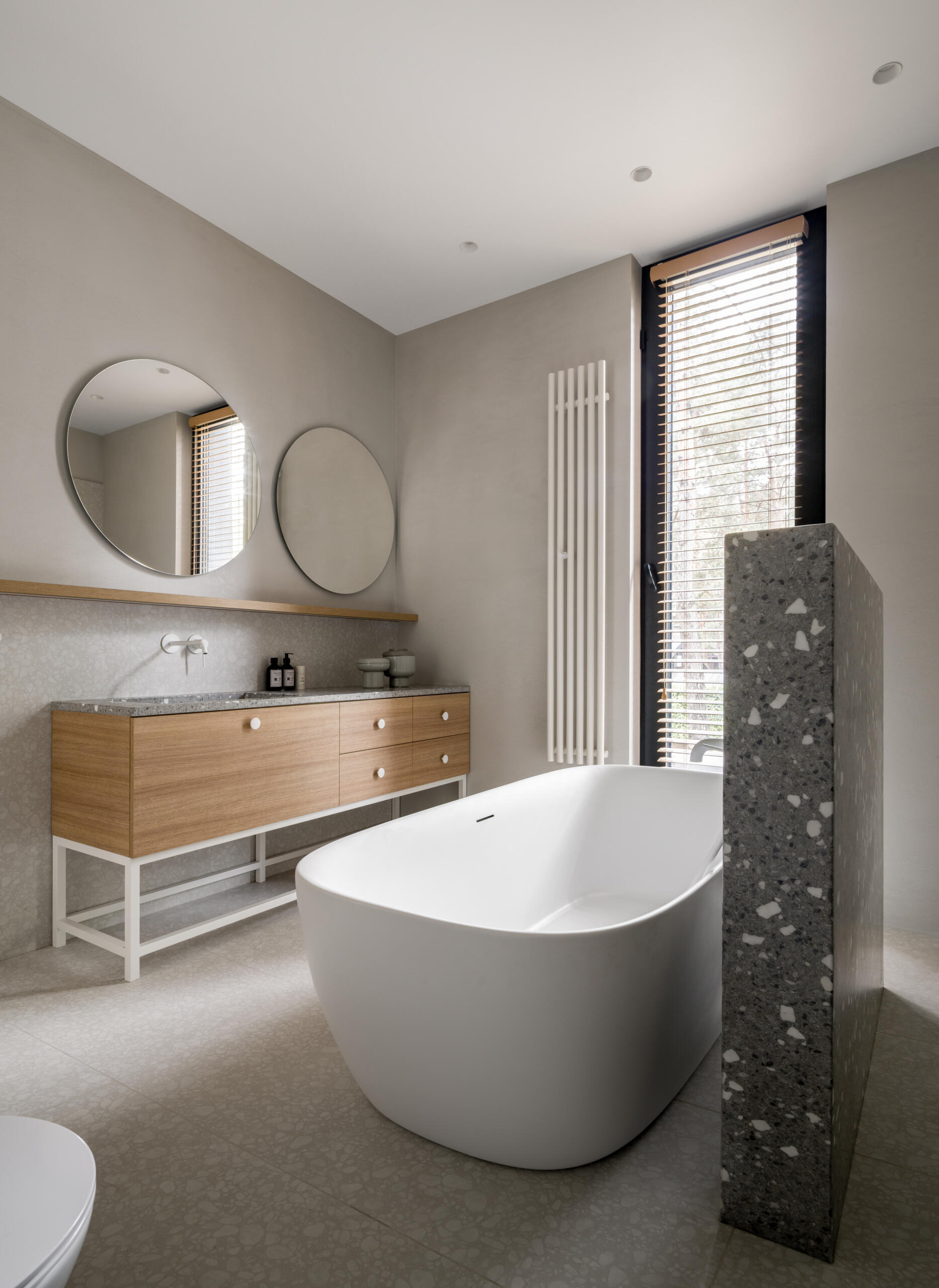 In this upstairs bathroom, soft greige walls and pale terrazzo-style tiles shape a quiet, balanced space centered around a sculptural white soaking tub. Daylight filters through wood blinds, warming the oak vanity and drawing attention to the speckled stone countertop and partition.