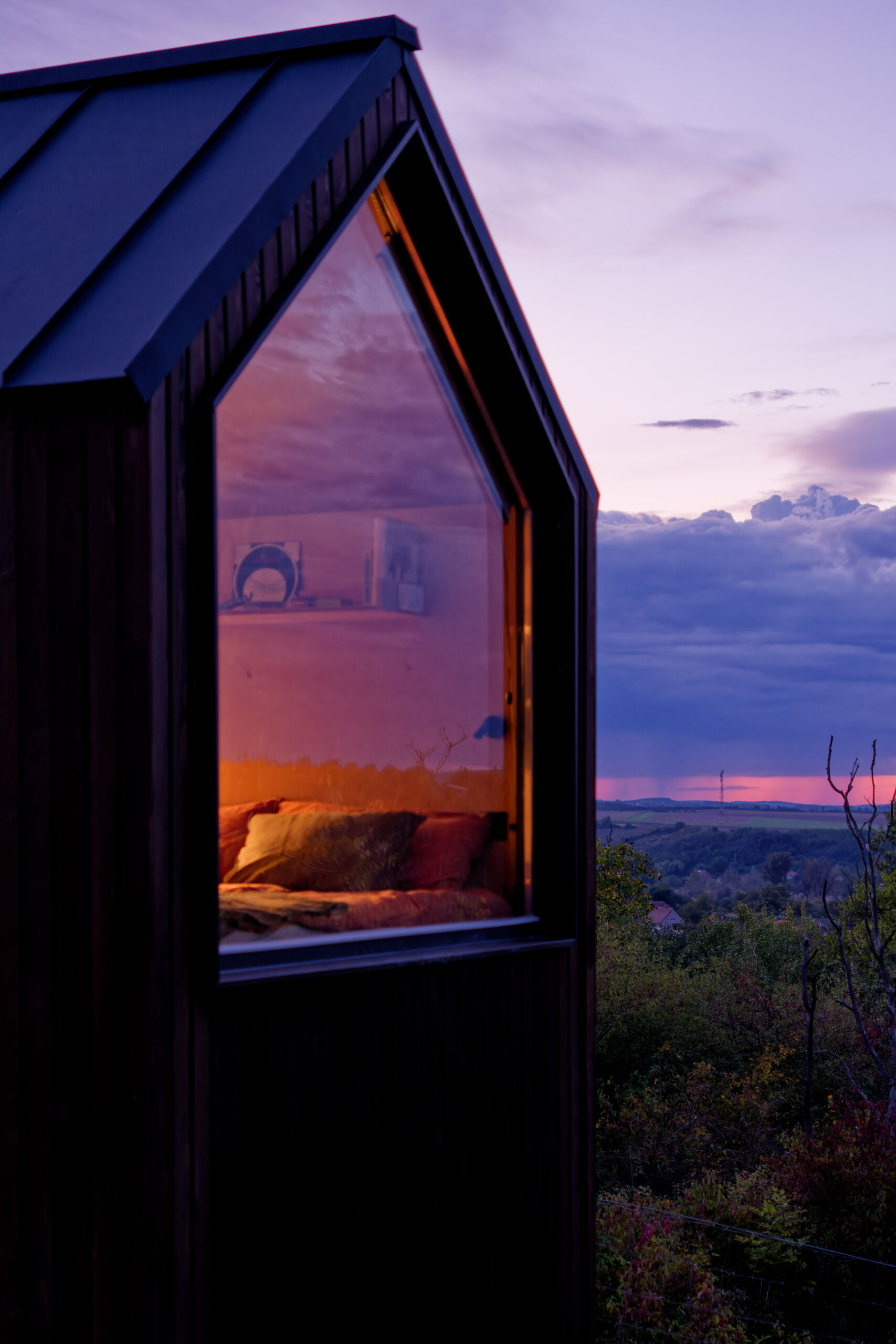 As night falls, the cabin glows softly above the village below. The illuminated trapezoidal window turns this small structure into a quiet landmark in the Hungarian hills, showing how thoughtful design transforms even the simplest form.