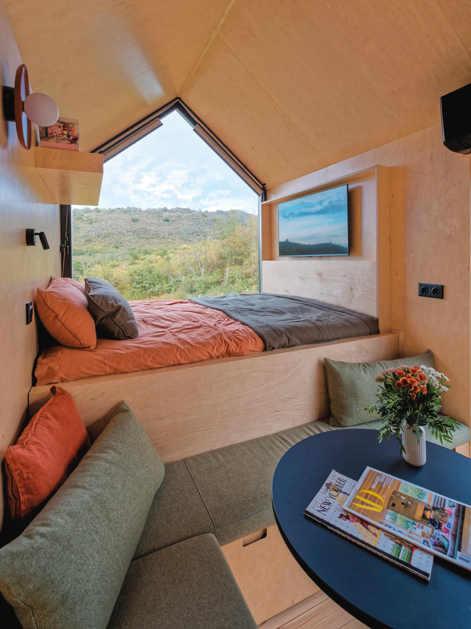 Light birch plywood, a visible gabled roof, and a striking trapezoidal window make this micro cabin interior feel surprisingly spacious. Included in the cabin is a sleeping area, lounge, kitchen, loft, and bathroom.