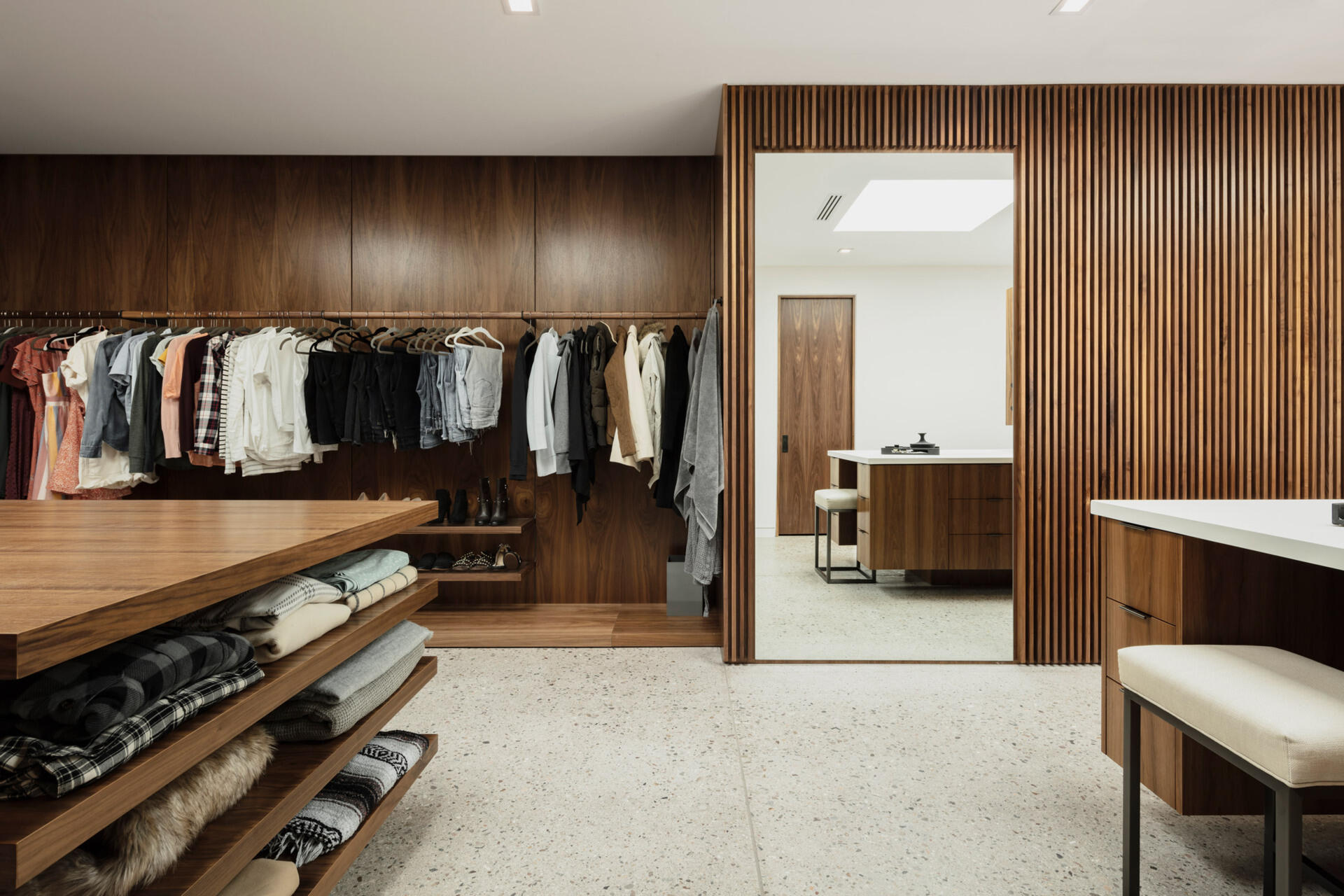 The primary suite unfolds into a spa-inspired bathroom with a skylight overhead. Dual vanities and a dedicated makeup area create generous space, while limestone surfaces and filtered light add a sense of calm. The vanity connects directly to a retail-inspired closet designed like a boutique, complete with racks, drawers, and a central table for additional storage.