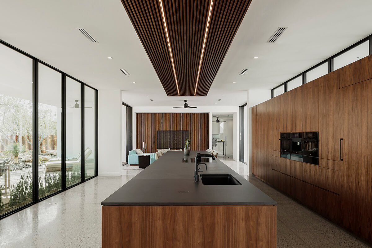 Light blue furnishings, walnut acoustic slats, and warm wood cabinetry define the open living space. A long island anchors the kitchen, with views stretching toward the patio and pool.