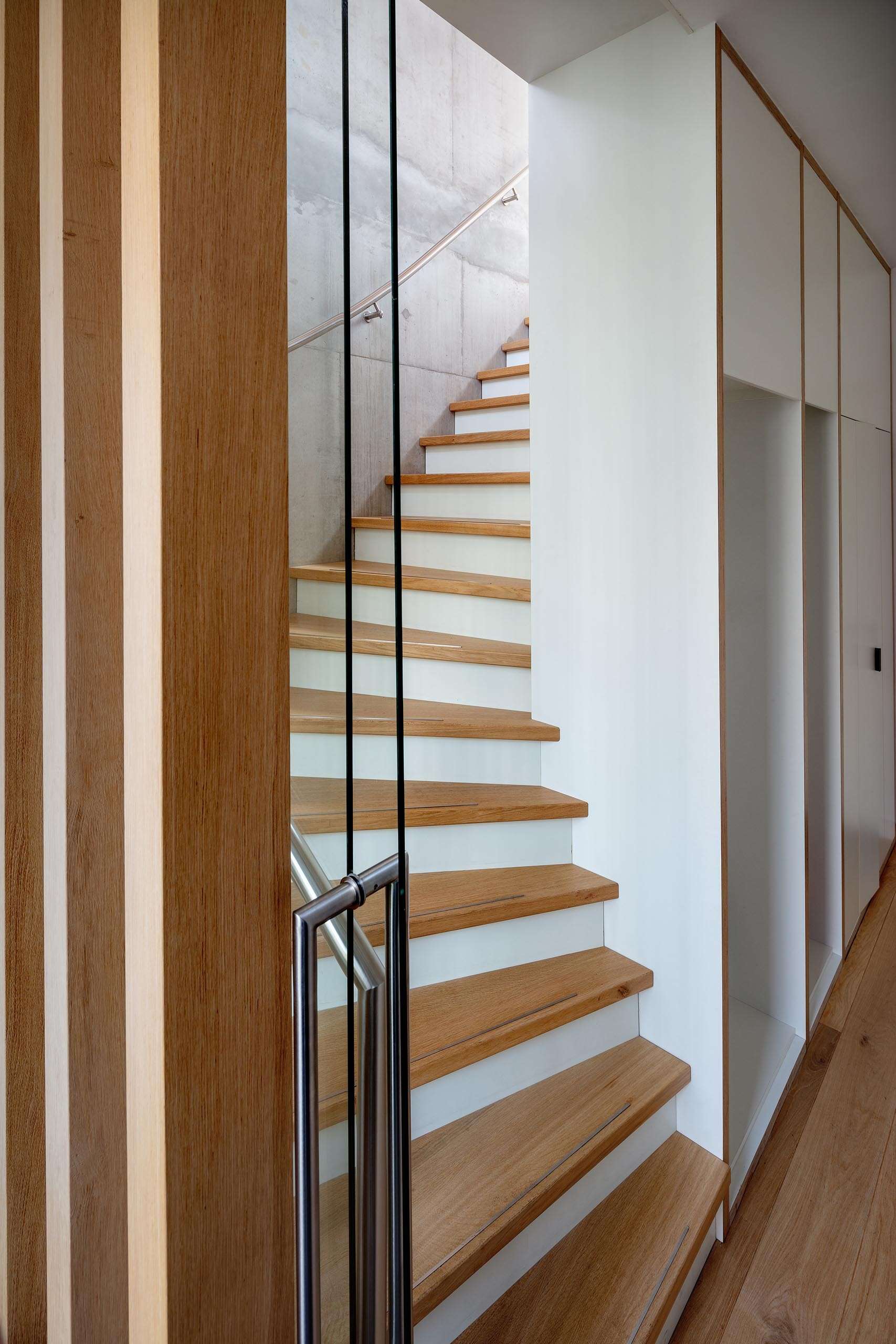 A central staircase with wood treads winds upward beside a full height custom bookshelf. Natural light from skylights and large windows keeps the vertical core bright and airy.