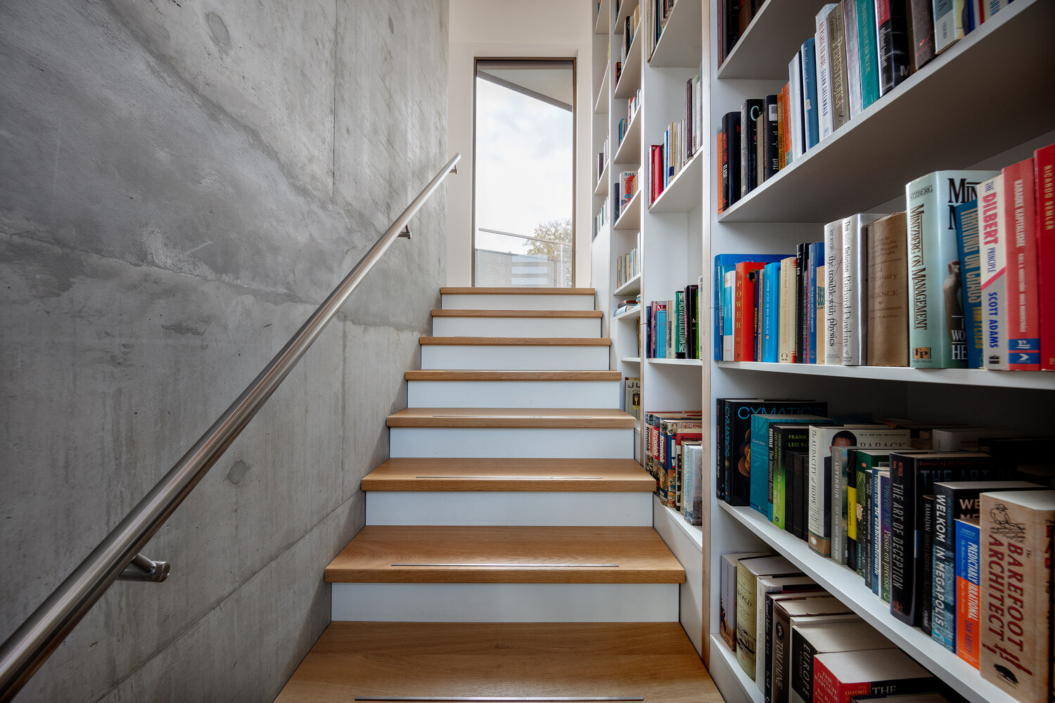 A central staircase with wood treads winds upward beside a full height custom bookshelf. Natural light from skylights and large windows keeps the vertical core bright and airy.