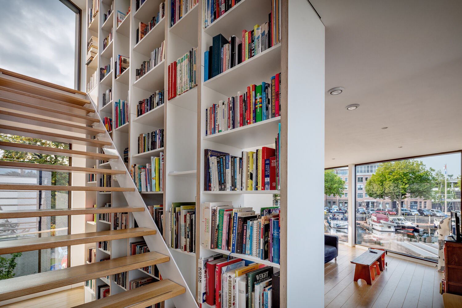A central staircase with wood treads winds upward beside a full height custom bookshelf. Natural light from skylights and large windows keeps the vertical core bright and airy.