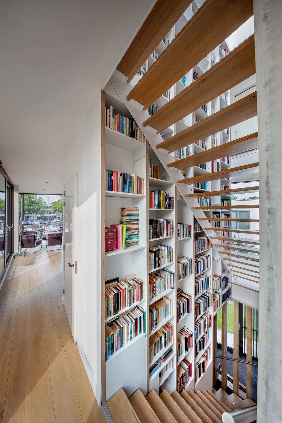 A central staircase with wood treads winds upward beside a full height custom bookshelf. Natural light from skylights and large windows keeps the vertical core bright and airy.