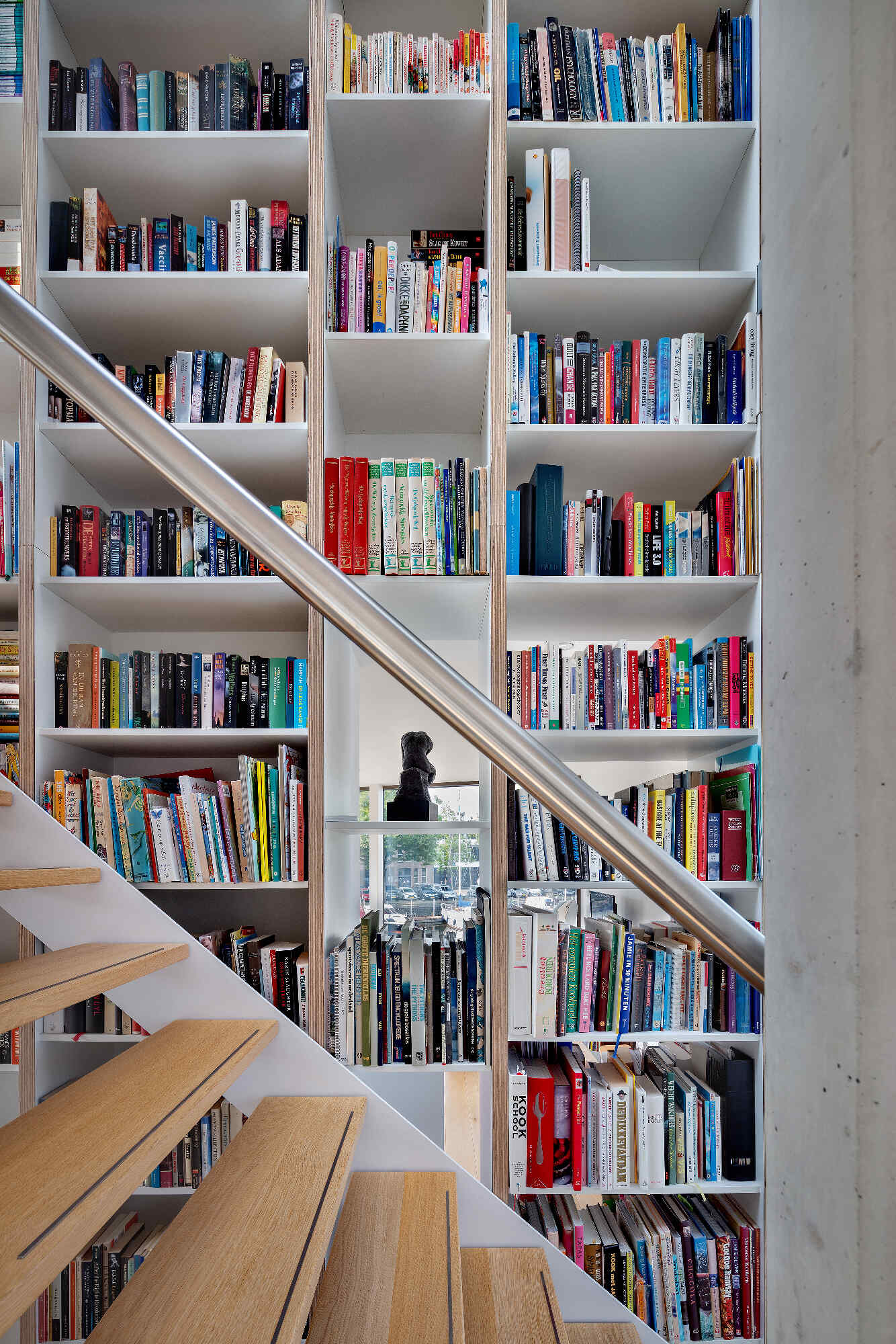 A central staircase with wood treads winds upward beside a full height custom bookshelf. Natural light from skylights and large windows keeps the vertical core bright and airy.