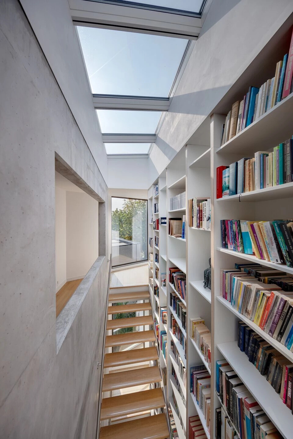 A central staircase with wood treads winds upward beside a full height custom bookshelf. Natural light from skylights and large windows keeps the vertical core bright and airy.